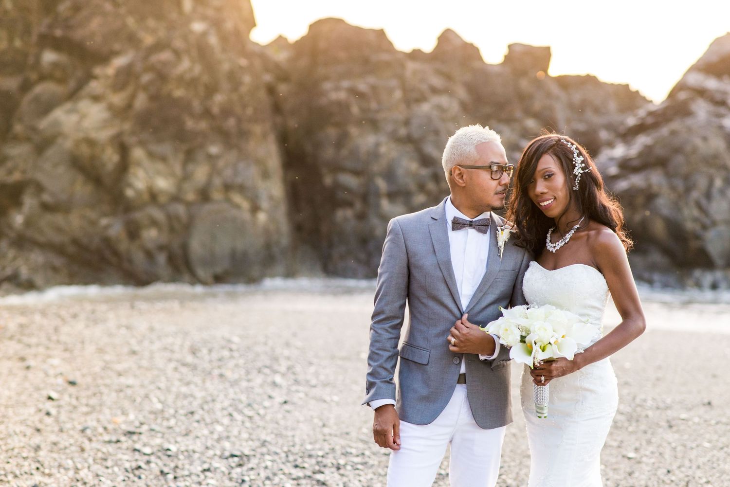Elegant interracial couple in wedding attire posing on a rocky beach at golden hour, bride holding white bouquet.
