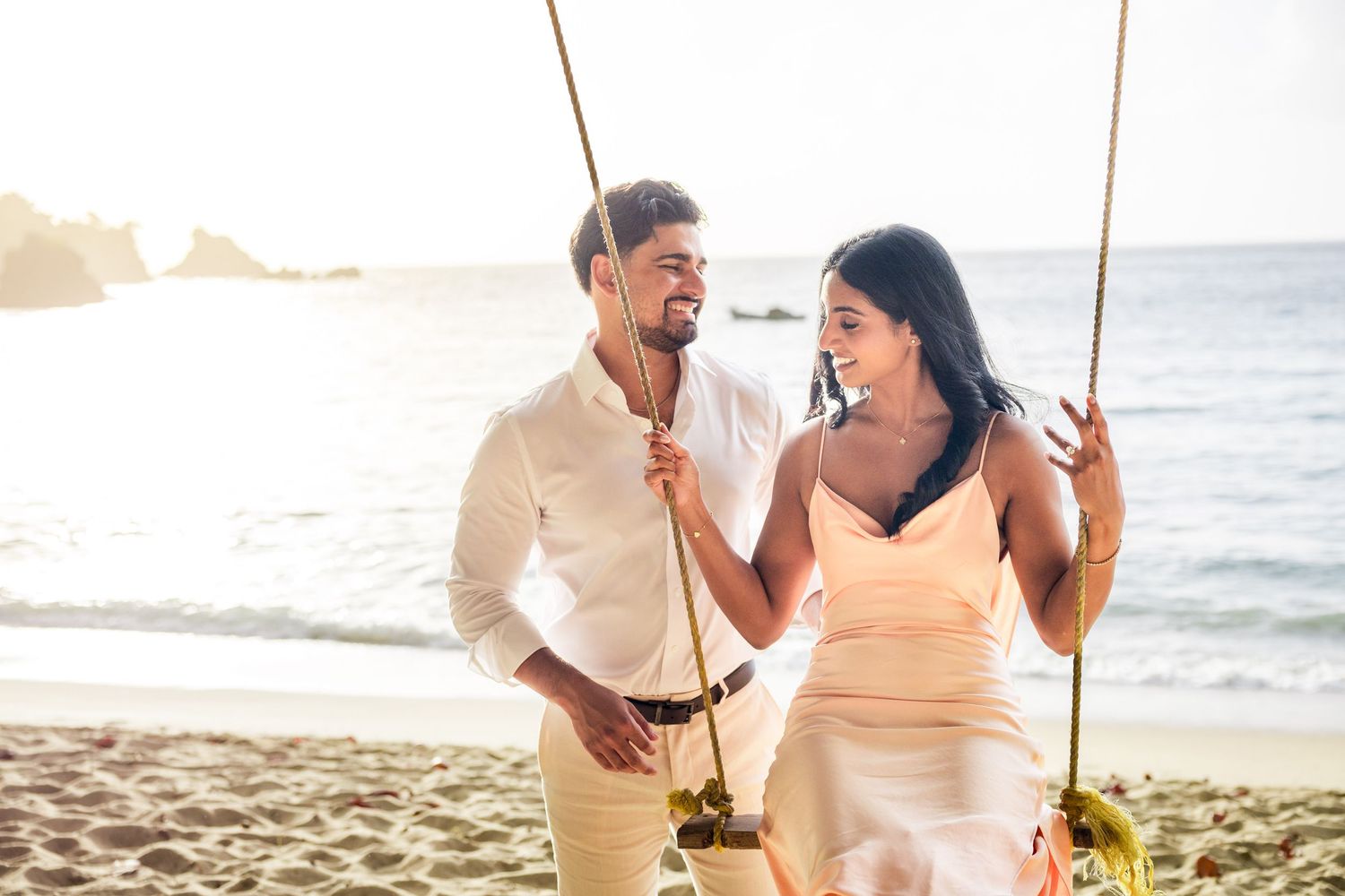 Couple smiling at each other on beach swings at sunset, man in white shirt and woman in yellow dress.