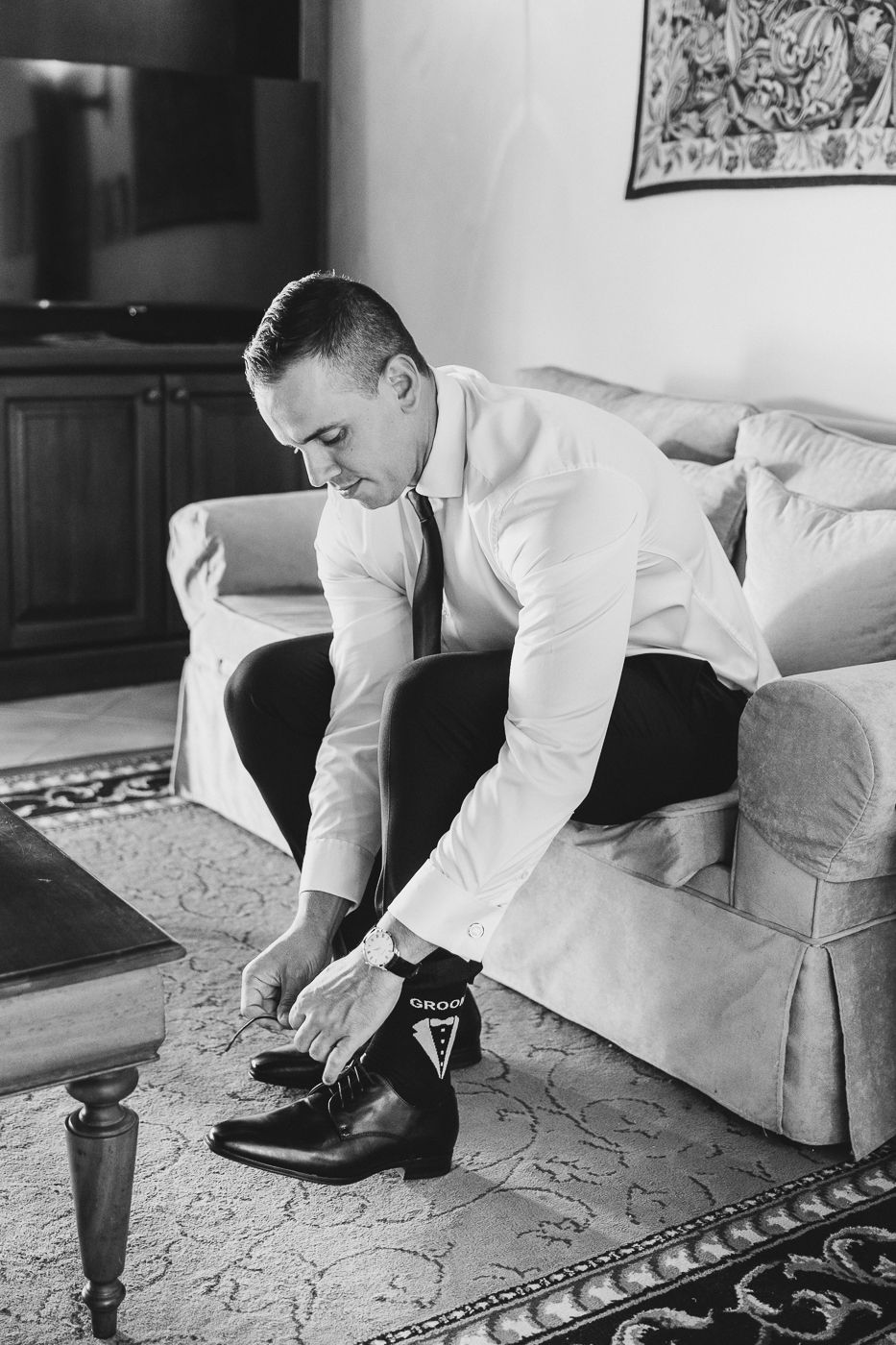 Groom in white shirt putting on dress shoes while seated on a sofa in black and white photo.