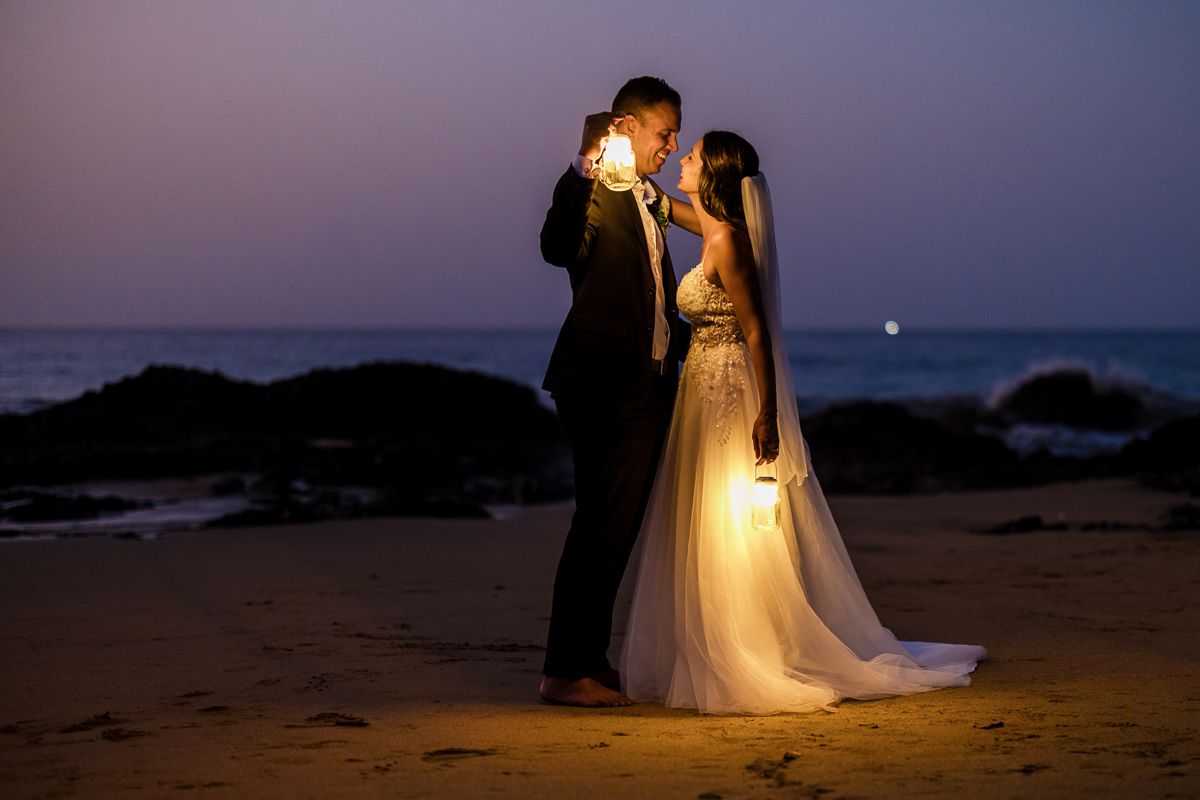 Bride and groom share a romantic moment on a beach at twilight, holding glowing sparklers during their wedding celebration.