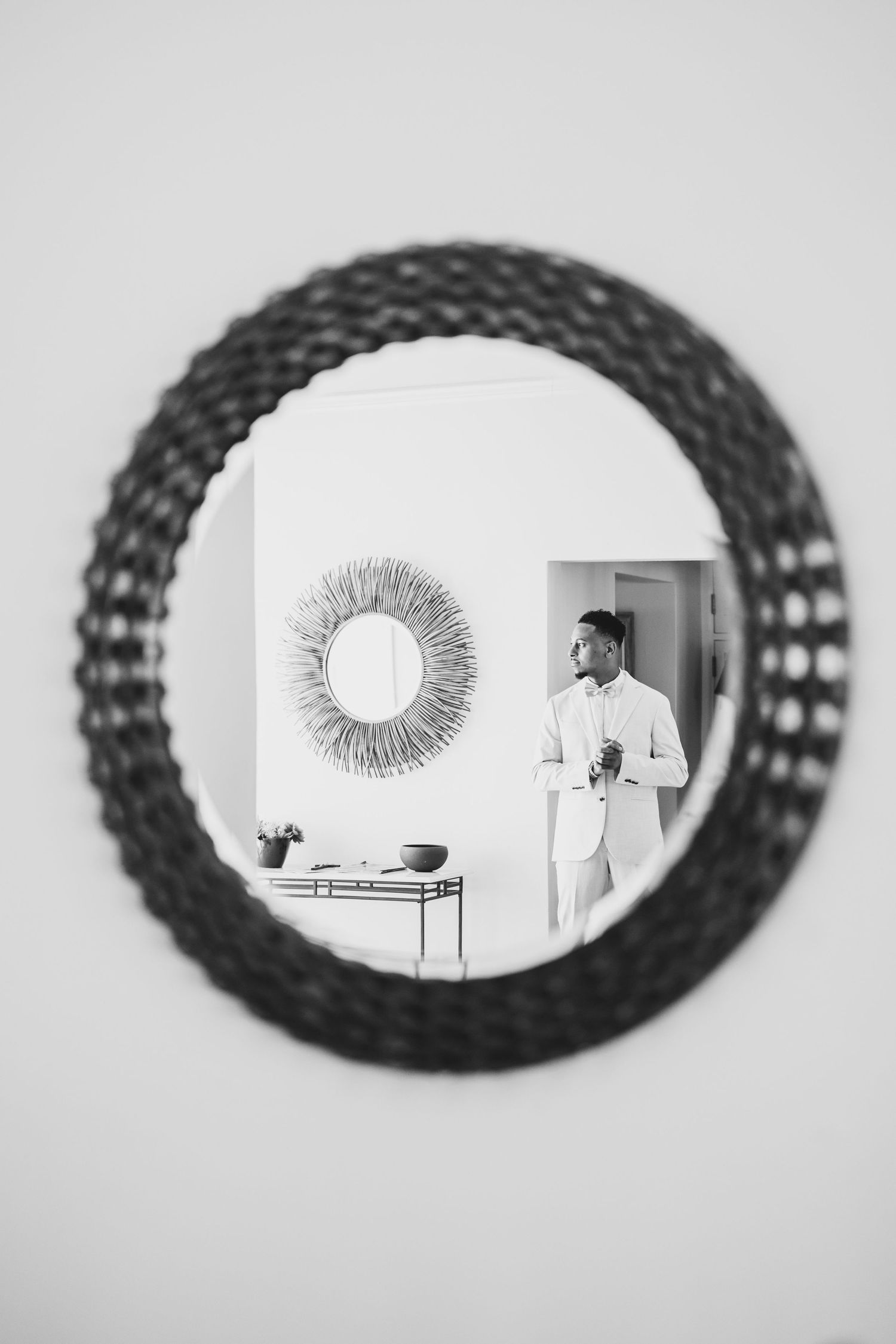 Black and white photo of a man in white suit reflected in a round woven mirror, with a sunburst mirror visible behind him.