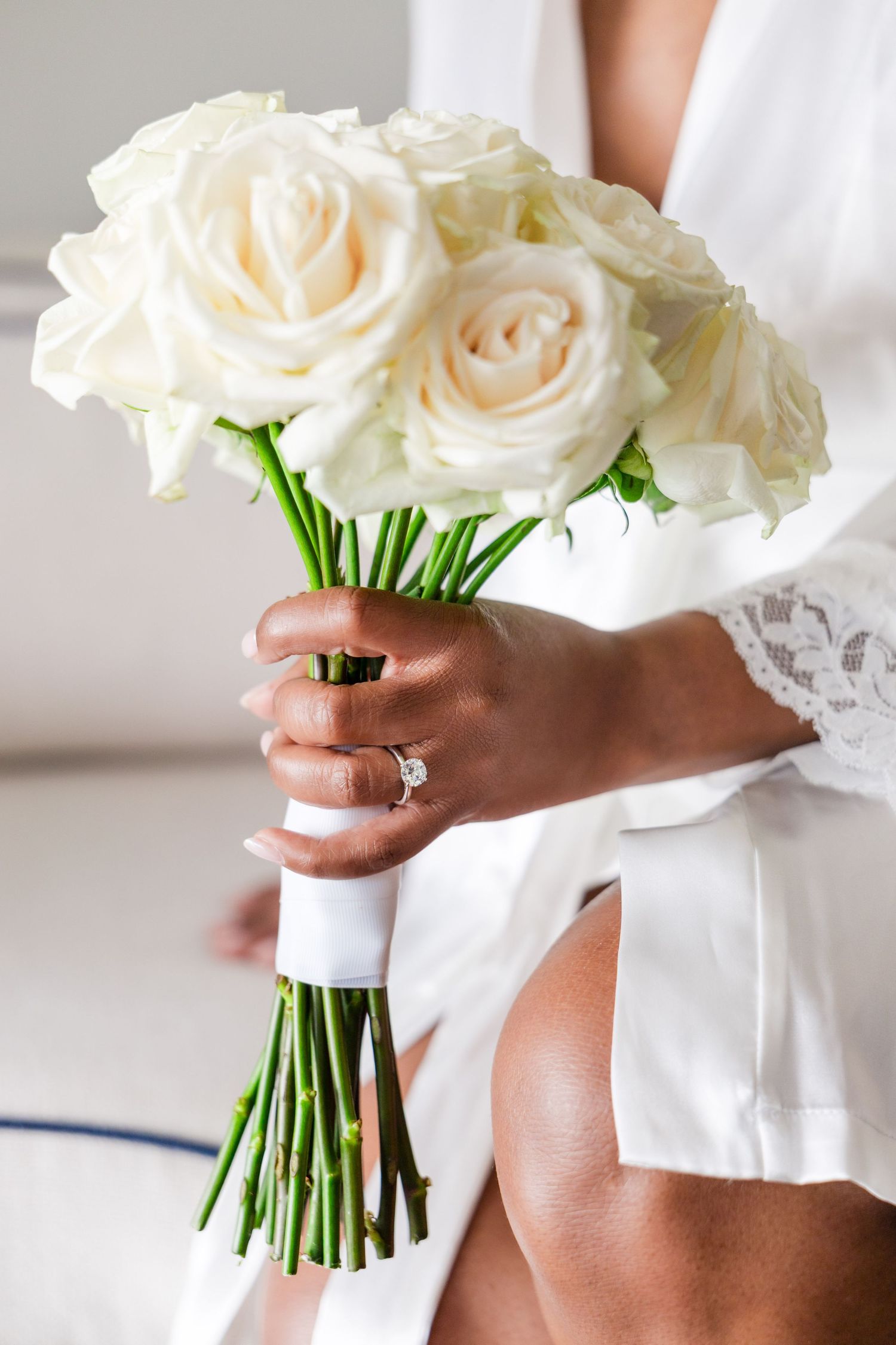 Bride holding elegant white rose wedding bouquet wrapped in white ribbon, wearing lace robe and diamond engagement ring.