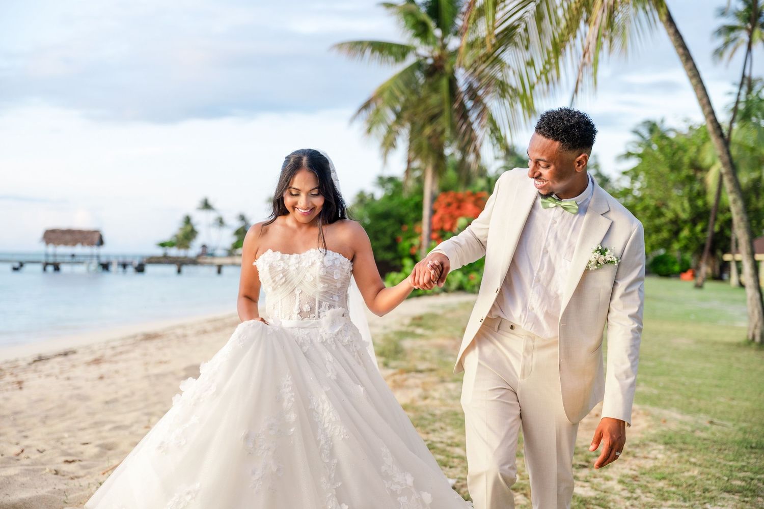 Bride in white ballgown and groom in beige suit walk hand in hand on a tropical beach with palm trees.