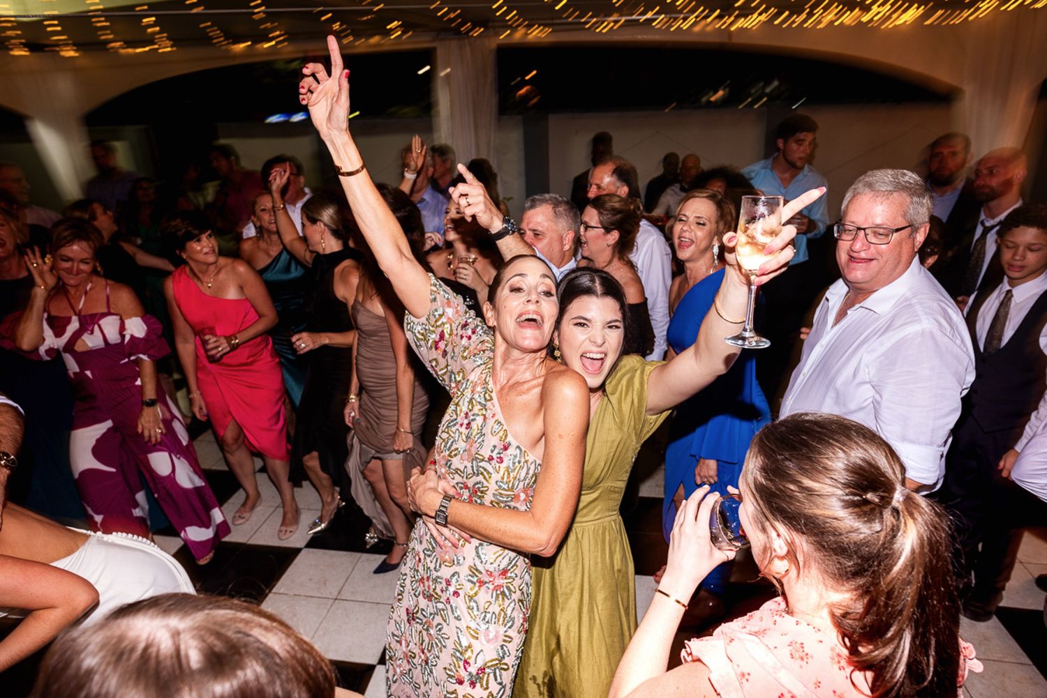 Wedding guests dance and celebrate on a crowded dance floor under warm lighting at a reception party.
