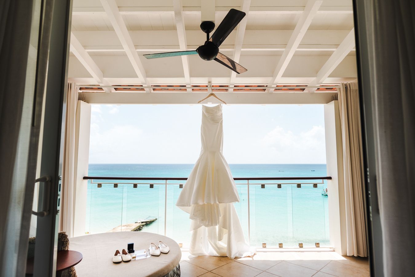 White wedding dress hangs in oceanfront room with ceiling fan and stunning beach view.