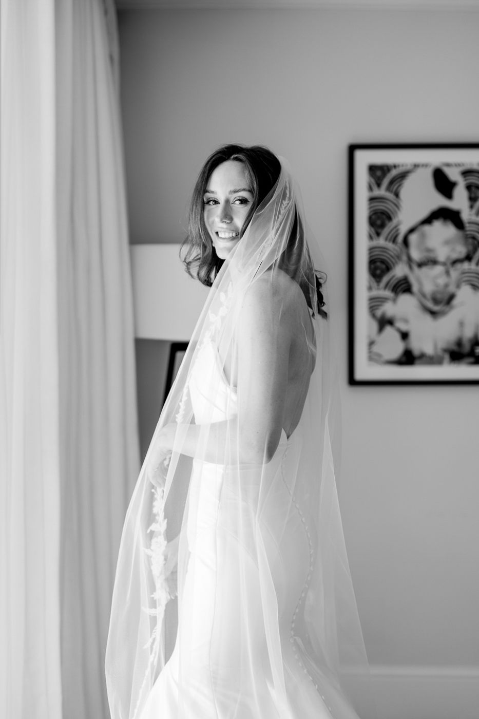 A bride in a flowing veil poses against a light wall with framed artwork.