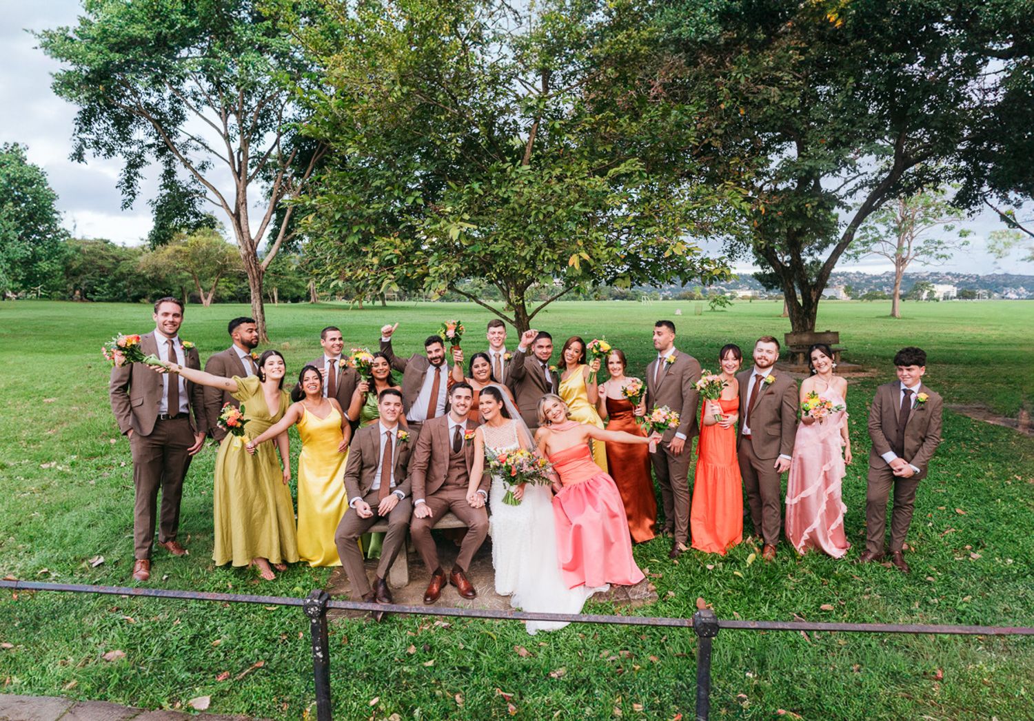 Wedding party members pose outdoors in vibrant yellow and coral dresses under trees at a green countryside venue.