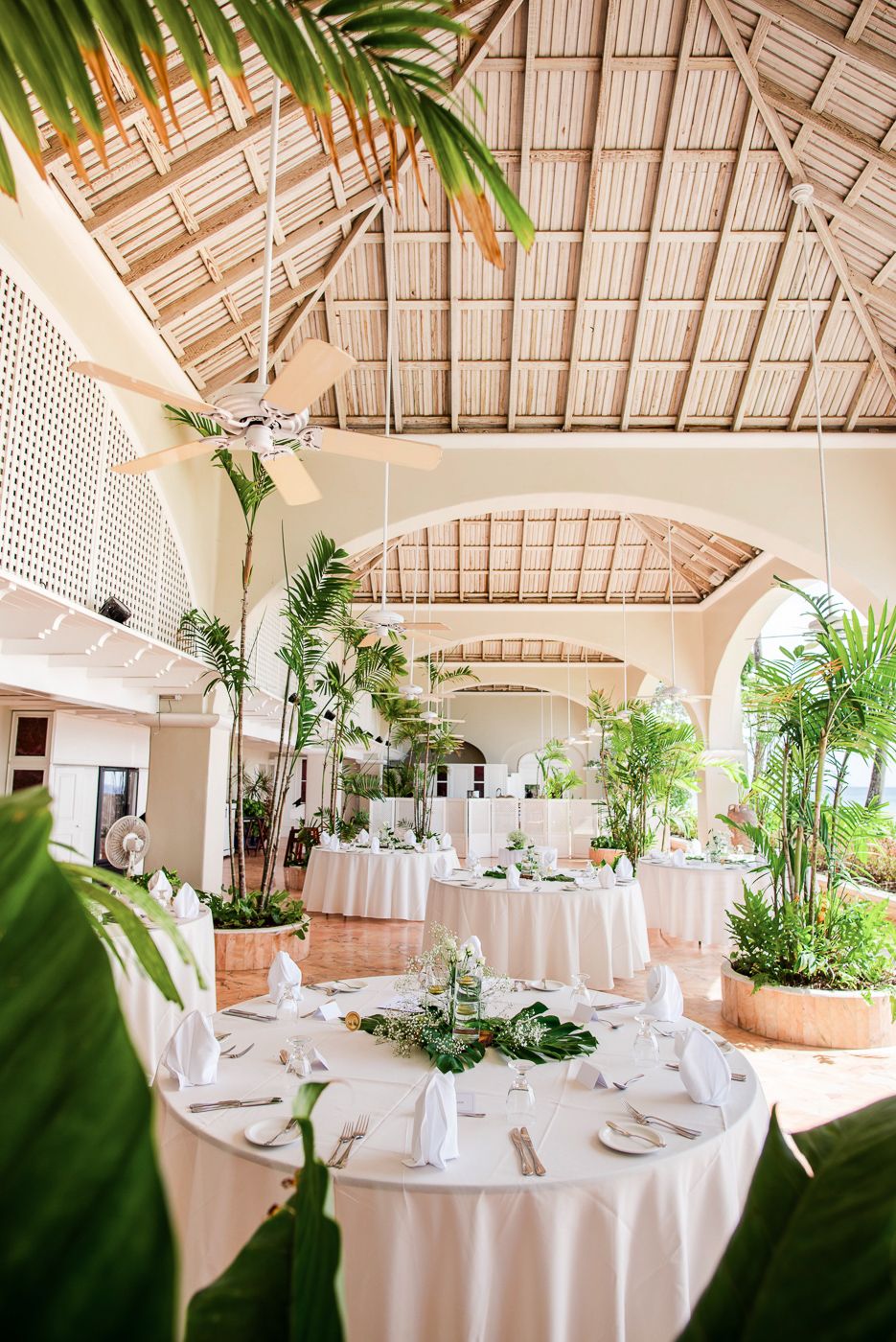 Spacious tropical dining room with vaulted bamboo ceiling, white tablecloths and potted palms.