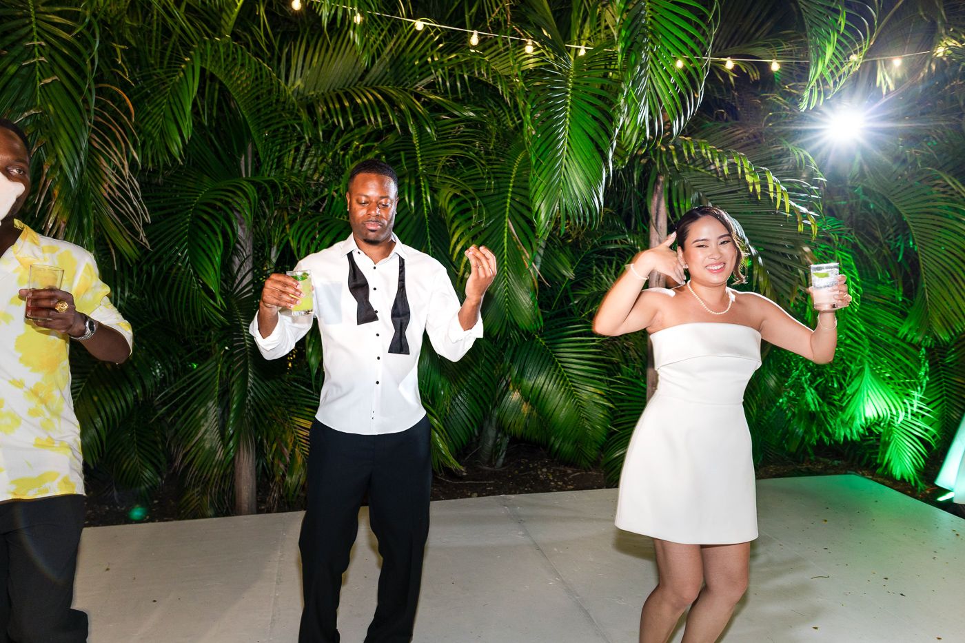 Wedding guests dance and celebrate under palm fronds and colorful party lighting.