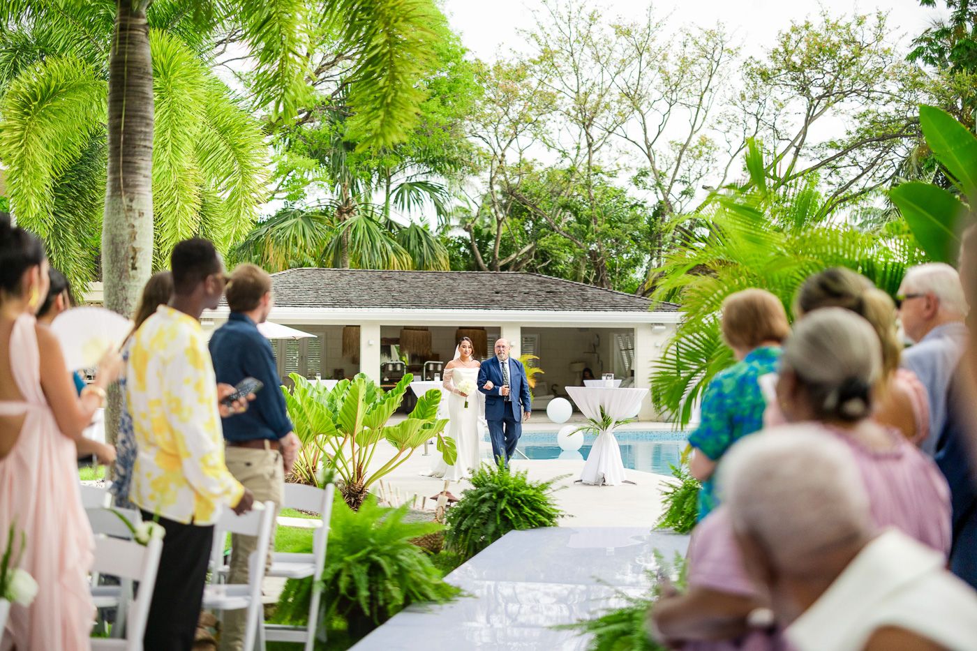 Outdoor wedding ceremony seating arrangement with white chairs and tropical landscaping.