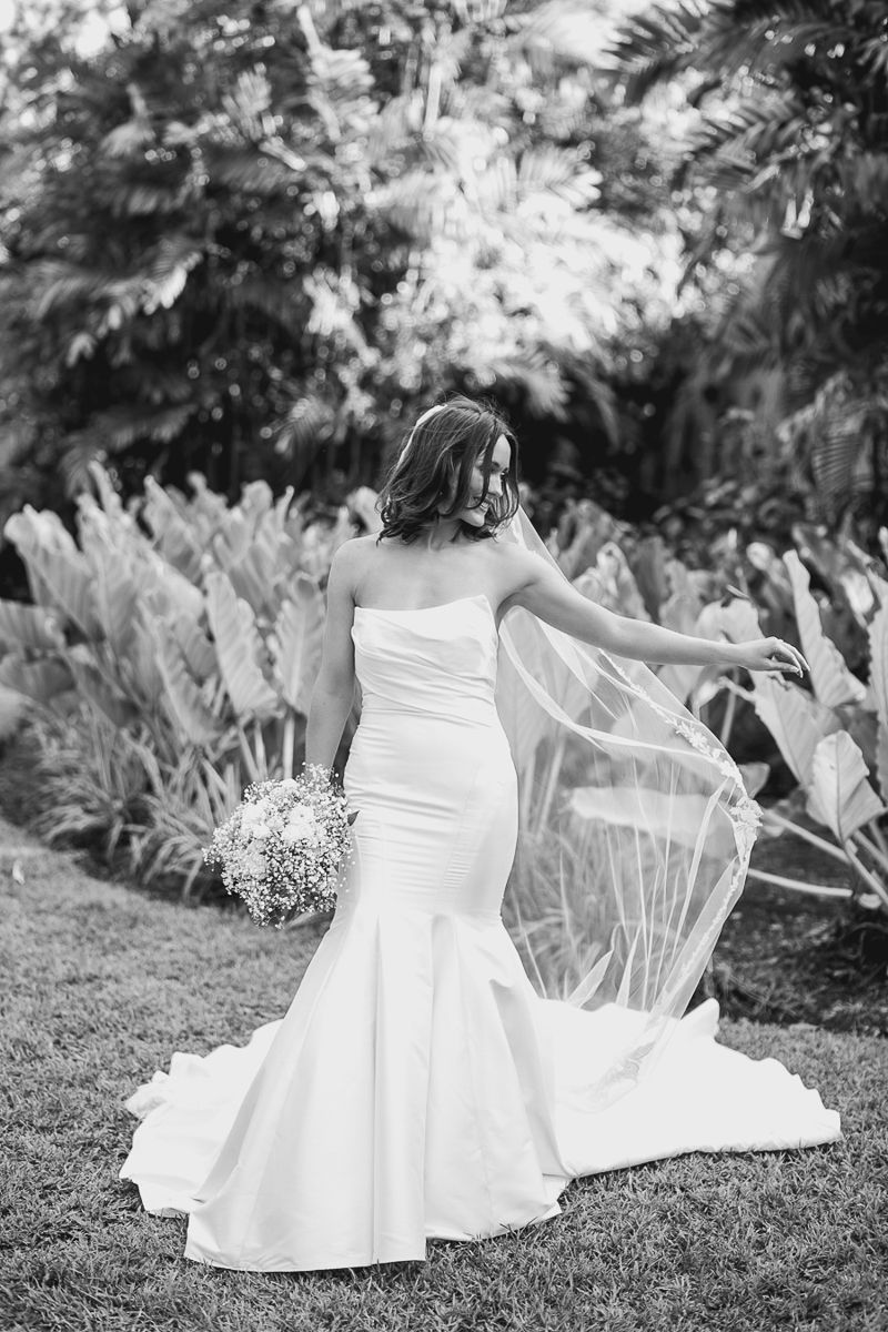 A white wedding dress with a flowing train laid out on grass with lush tropical foliage in the background.