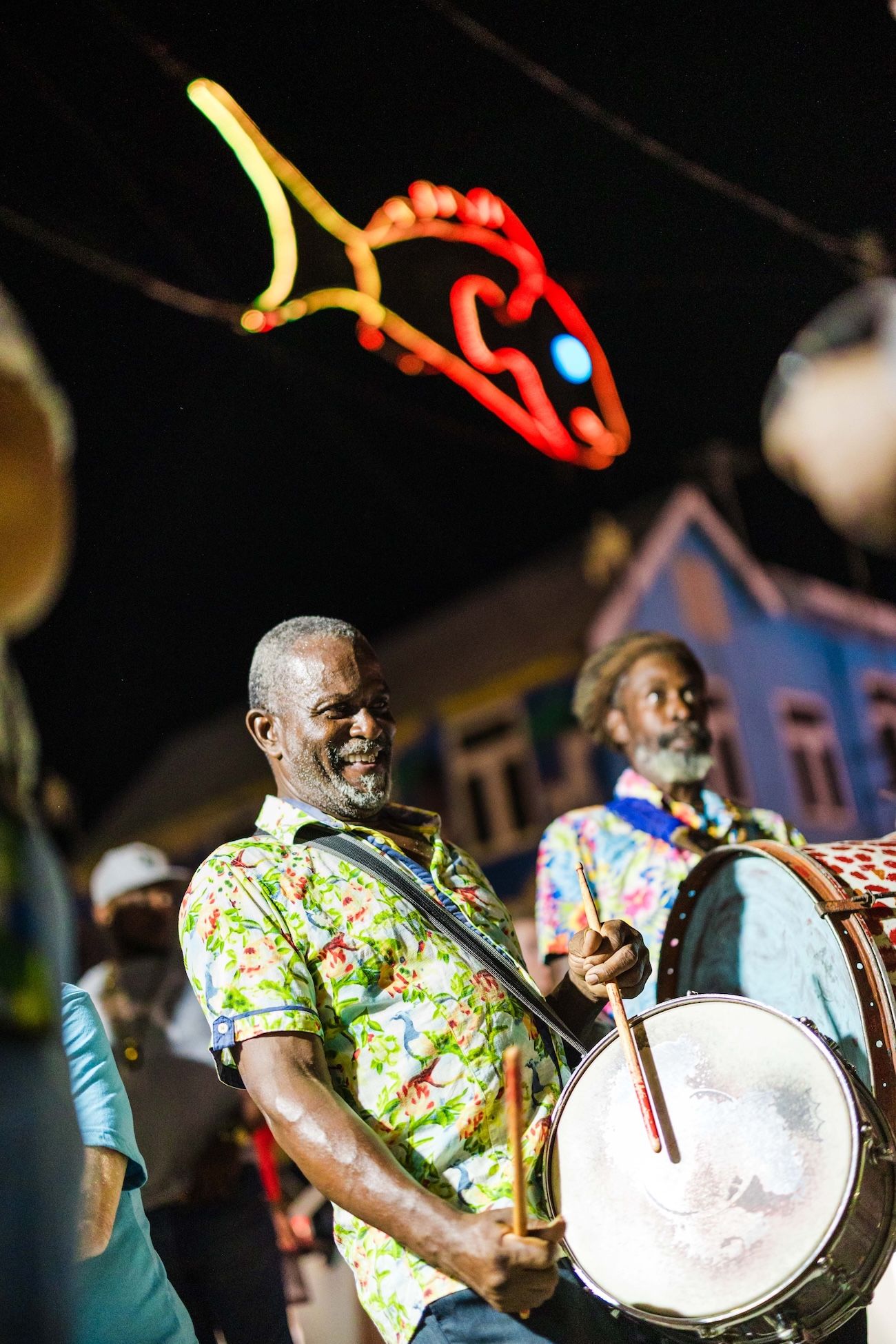 Smiling man playing bass drum in colorful floral shirt at vibrant night carnival parade with neon fish sign overhead.