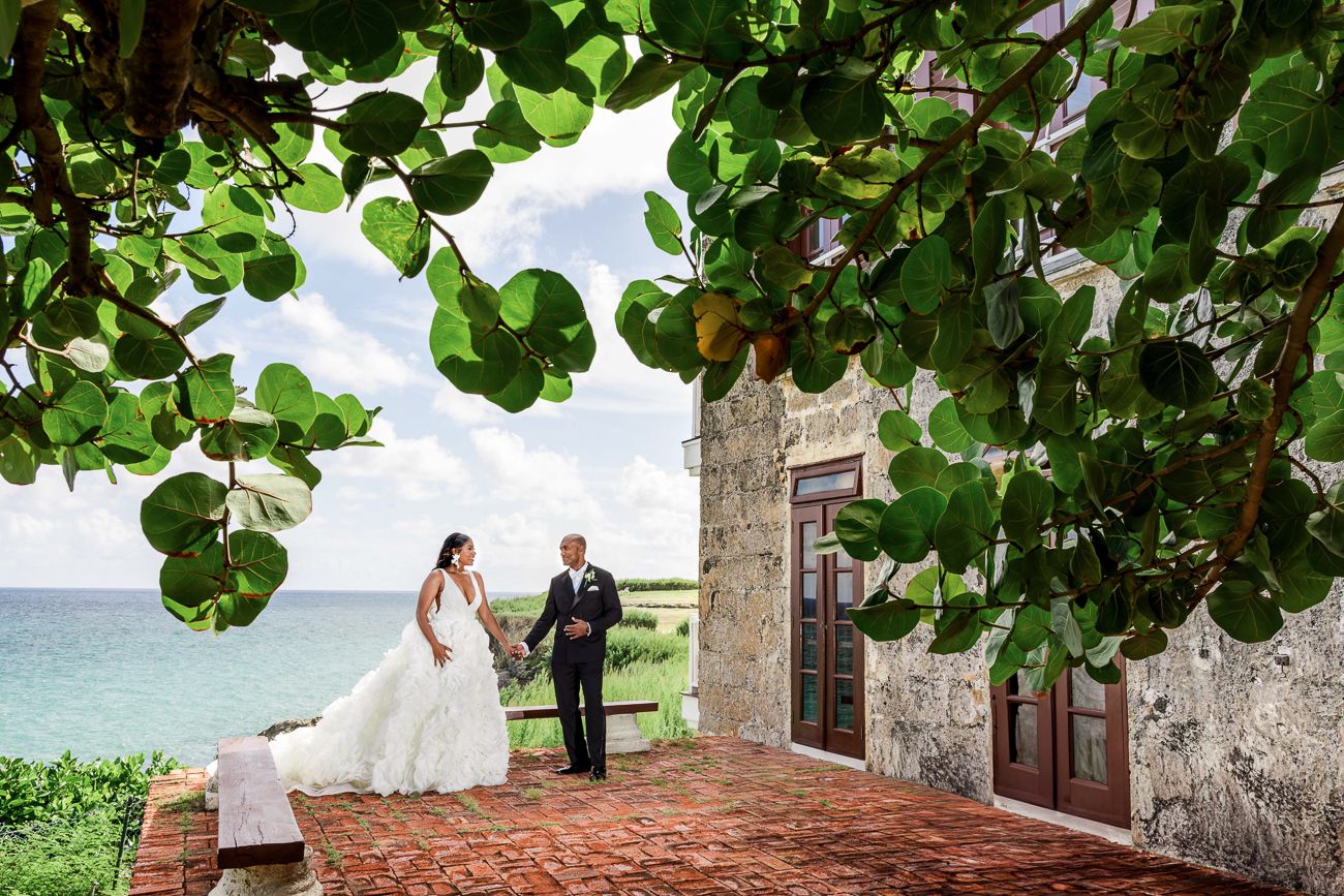 Bride and groom hold hands on a stone terrace overlooking the ocean, framed by lush green tropical leaves.