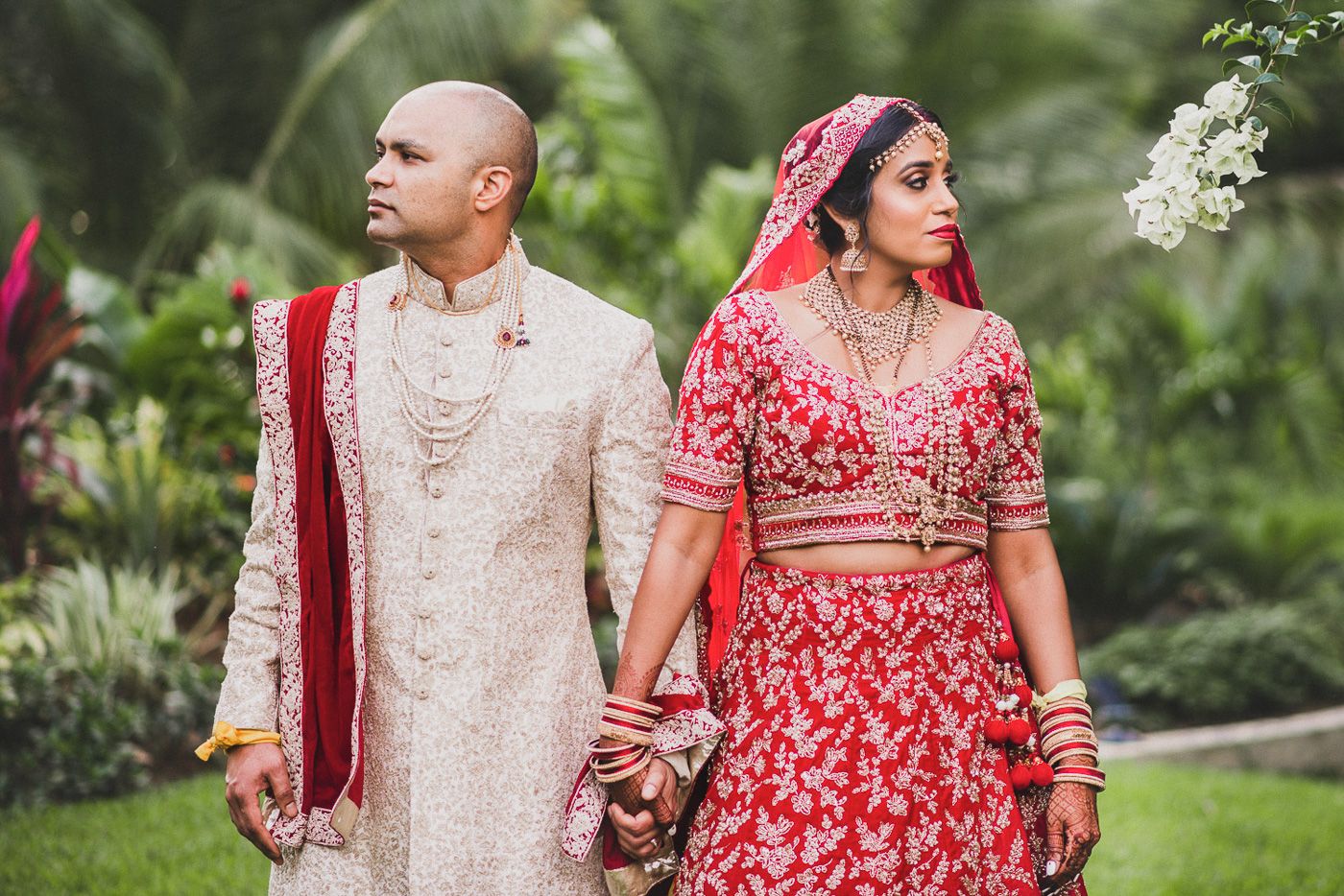 Indian bride in red lehenga and groom in cream sherwani hold hands during traditional Hindu wedding ceremony outdoors.