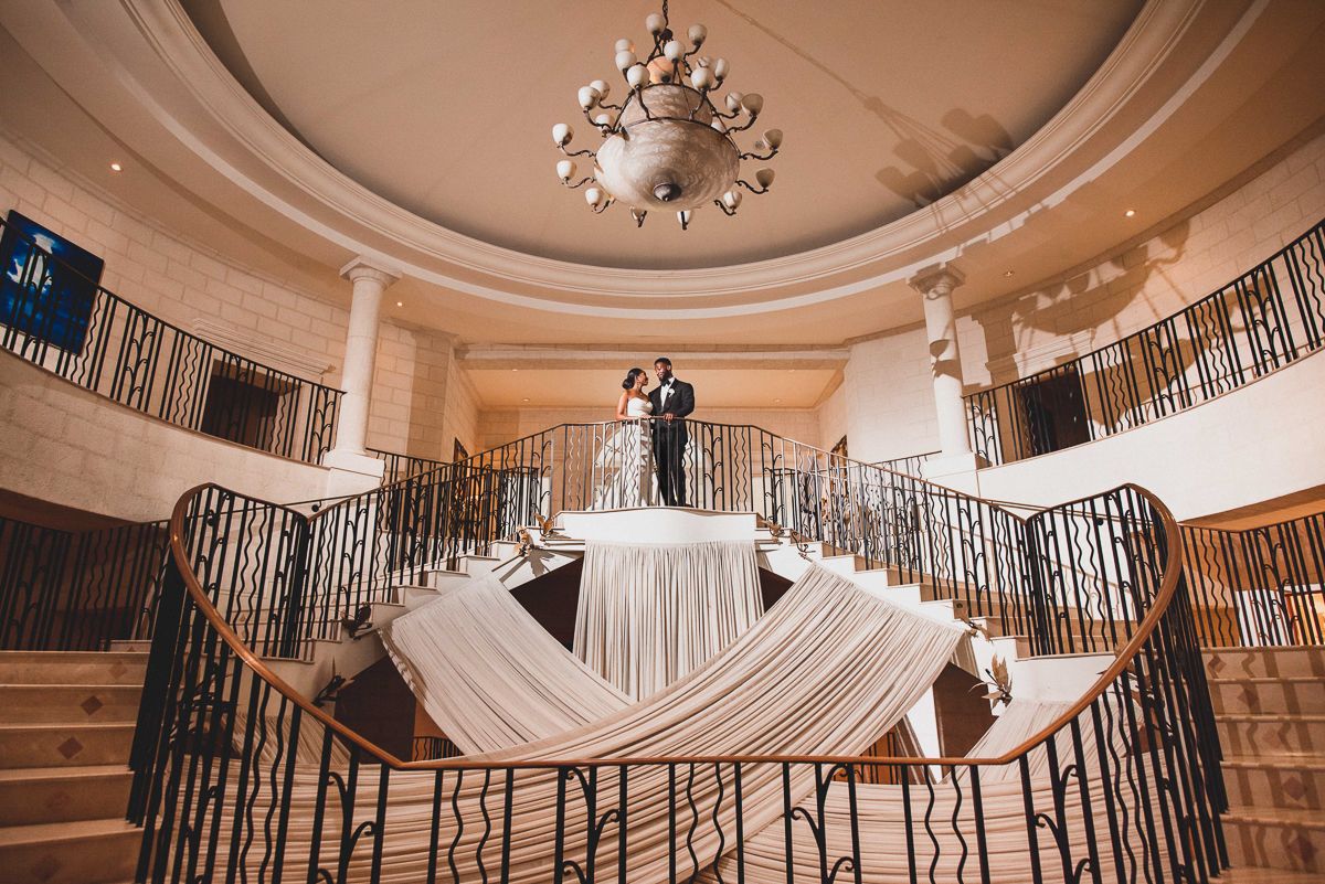 Bride and groom standing atop an elegant grand staircase with flowing white fabric in a luxurious wedding venue.