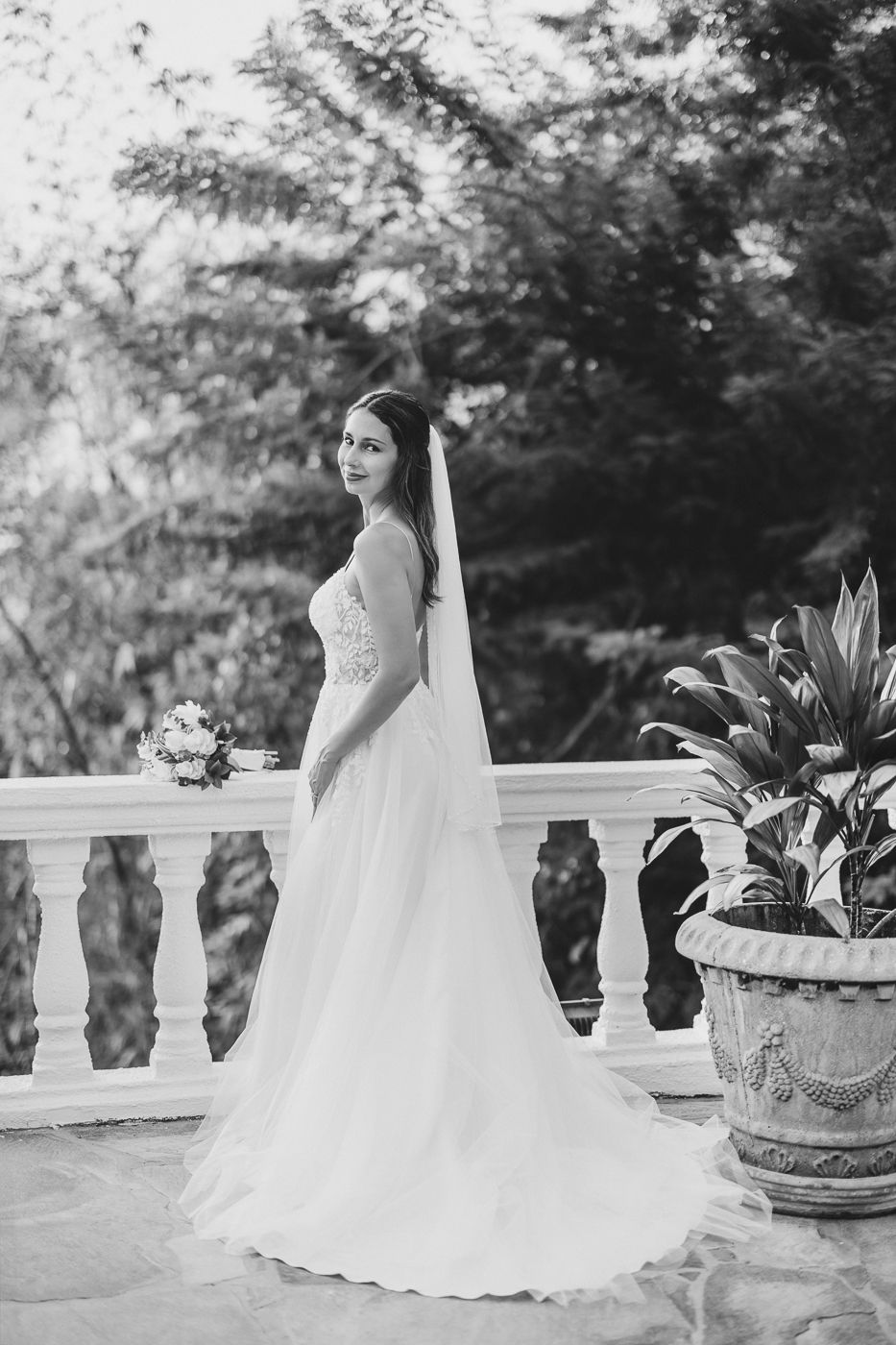 Elegant bride in a white lace gown and veil poses on a balcony surrounded by lush greenery.