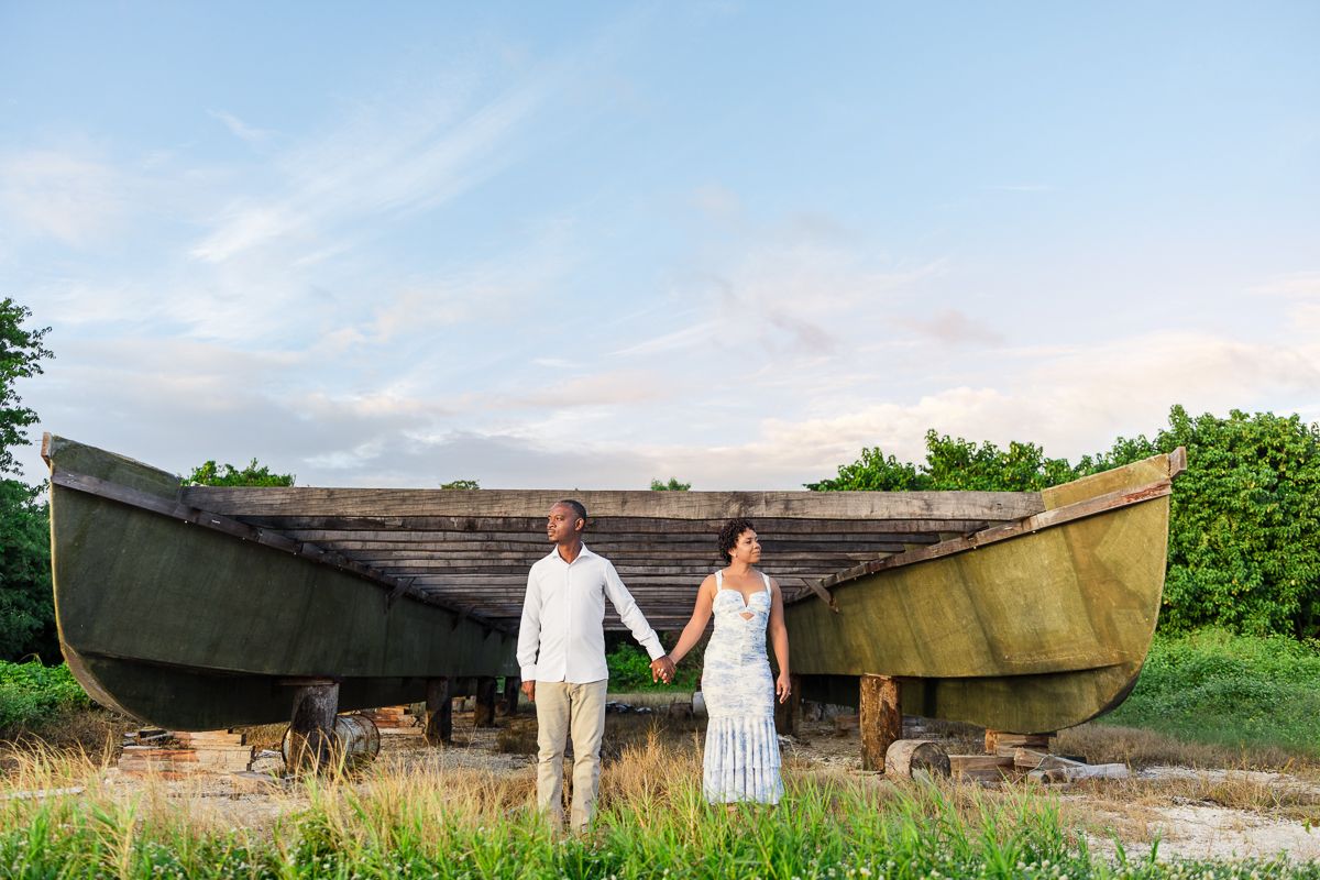 A couple poses together in a rustic outdoor setting with vintage wooden boats and lush greenery in the background.