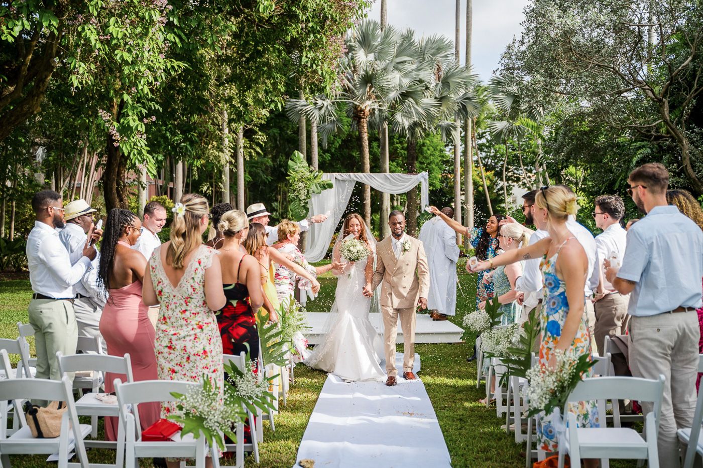 Wedding guests seated in white chairs during an outdoor ceremony surrounded by lush tropical gardens.