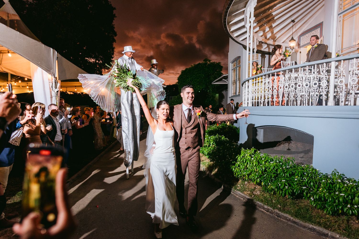 A couple exits their wedding venue at night while guests cheer and celebrate under dramatic evening skies.