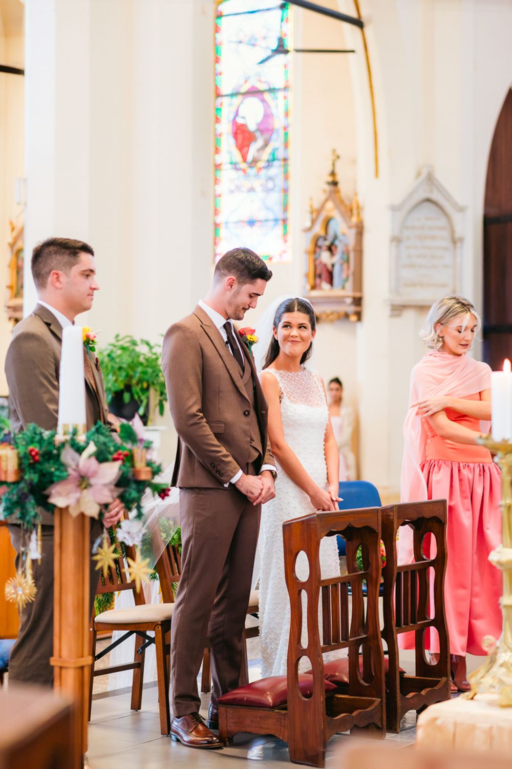 A row of wedding guests praying during a church ceremony with coral-colored bridesmaids' dresses visible.