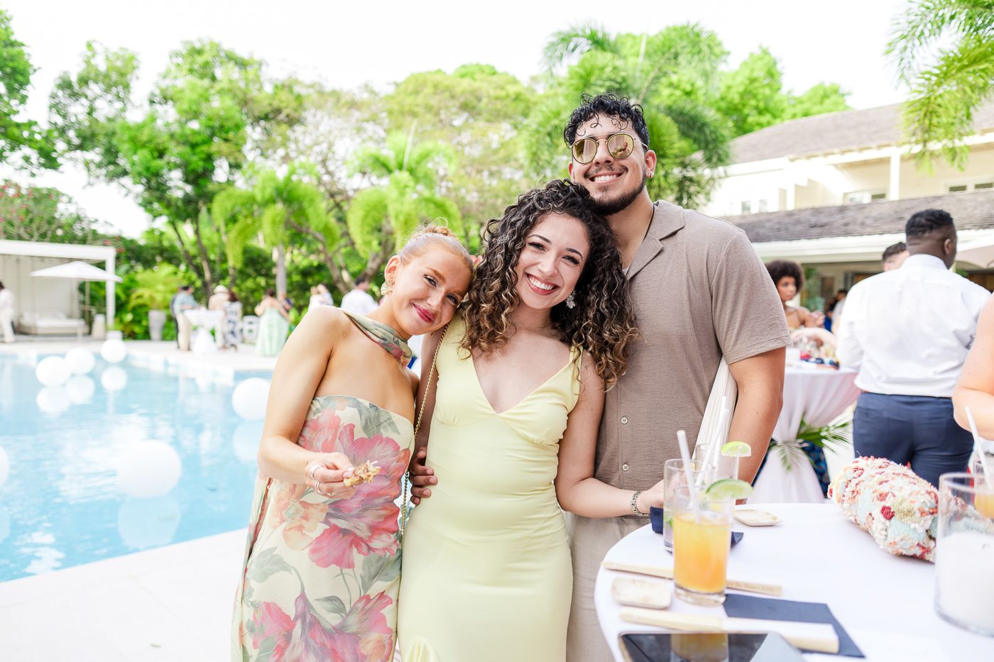 Three friends pose together by a pool during a celebration, sharing drinks and smiles.