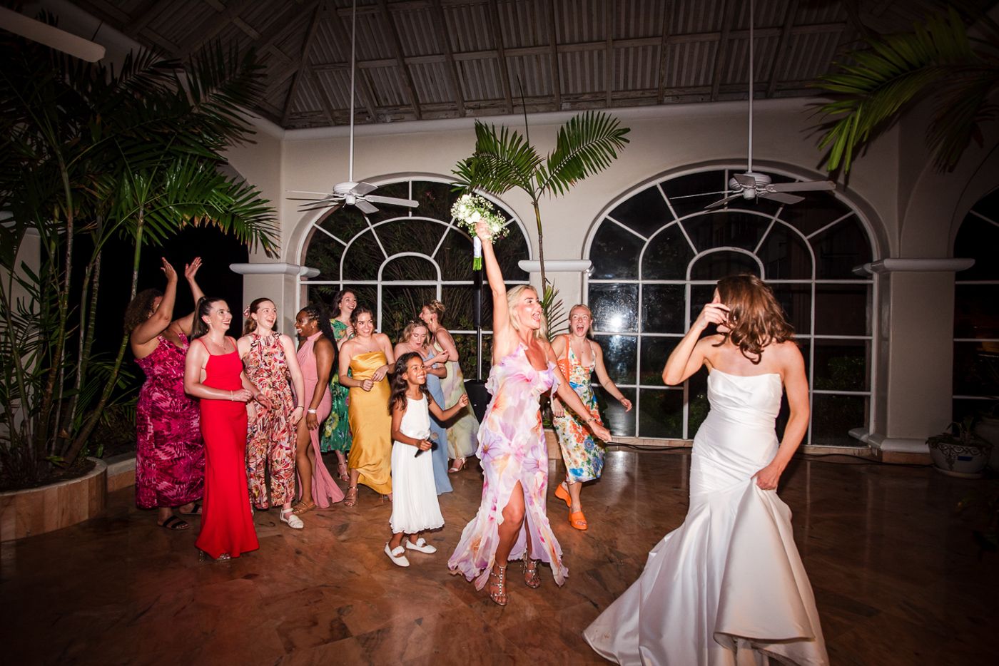 Wedding party dancing together in a tropical venue with arched windows and palm plants.