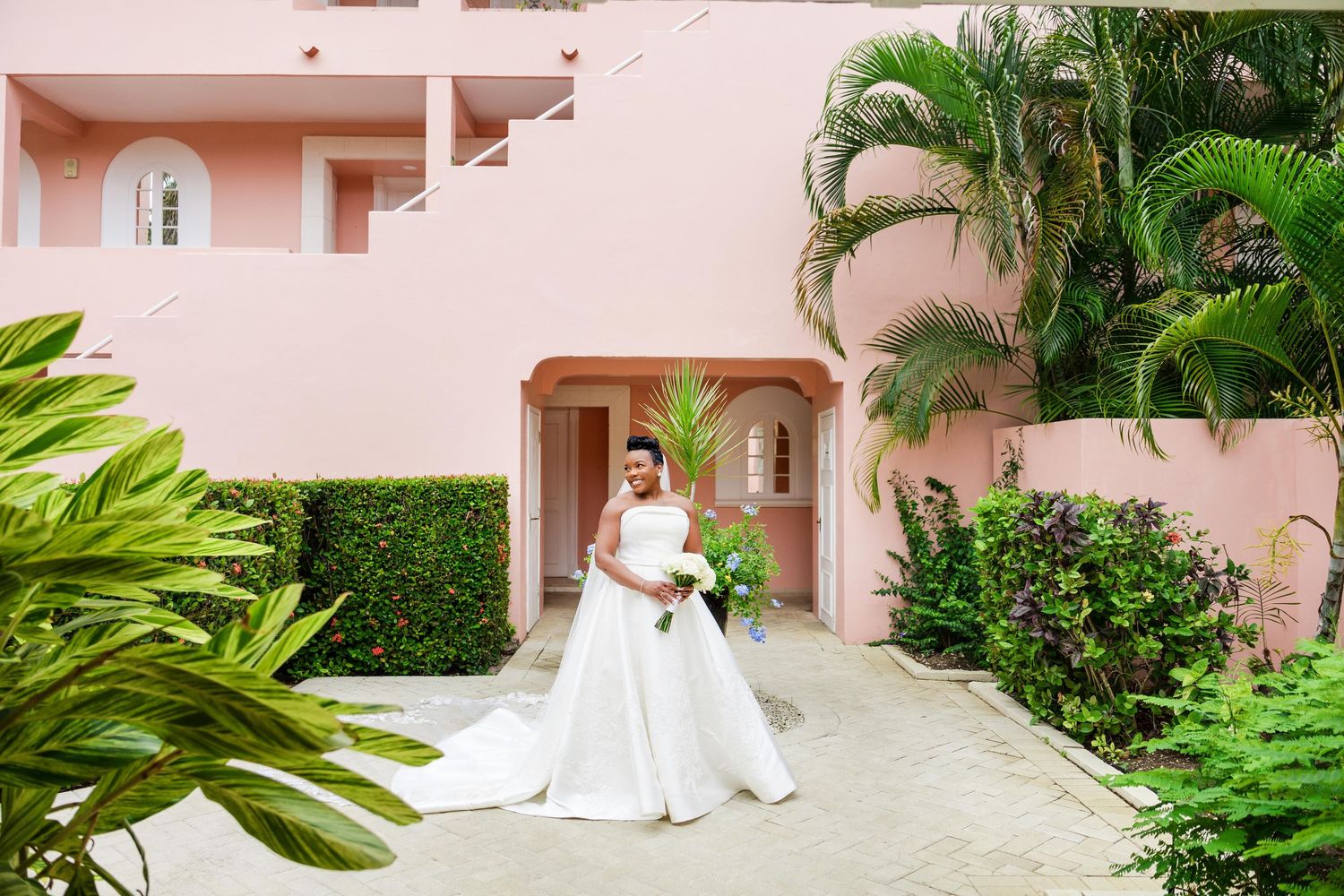 Bride in white ball gown poses in front of pink tropical resort building surrounded by lush green palm trees.
