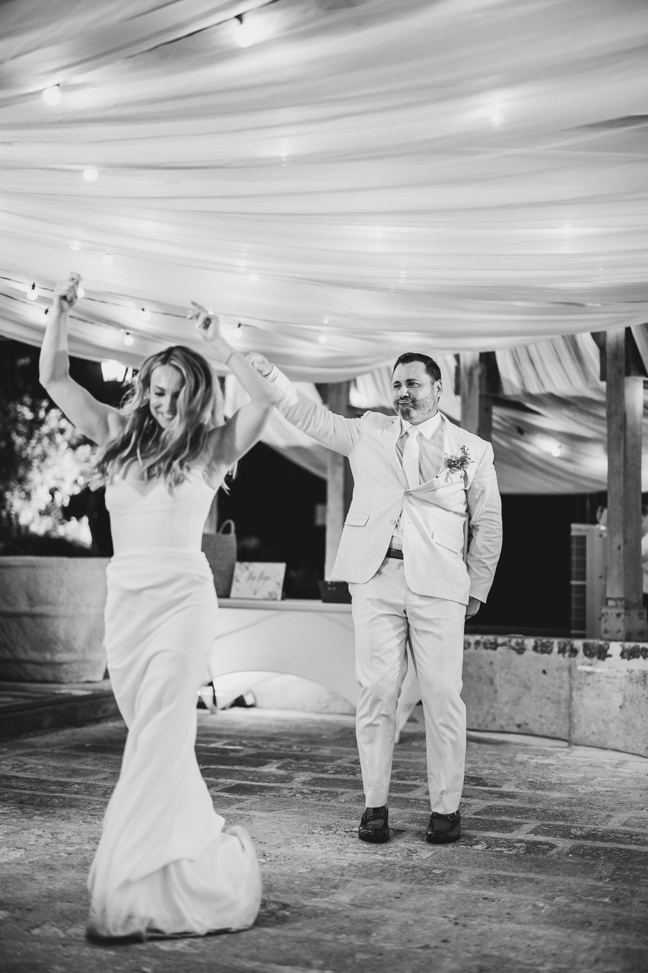 Black and white photo of bride and groom sharing first dance under draped fabric ceiling with string lights at wedding reception.