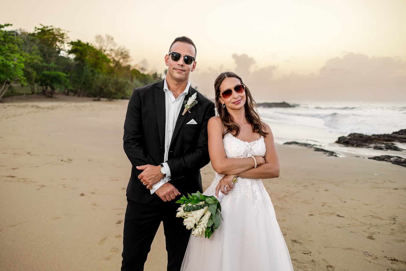 Bride and groom in sunglasses posing on a tropical beach, bride holding white bouquet in wedding attire.