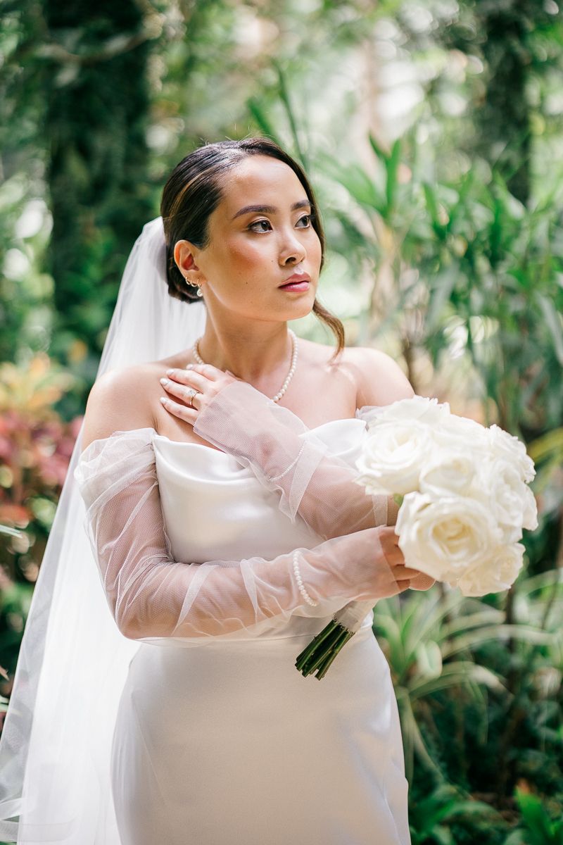 Elegant bride in off-shoulder white gown with sheer sleeves holds white rose bouquet, wearing veil in lush garden.