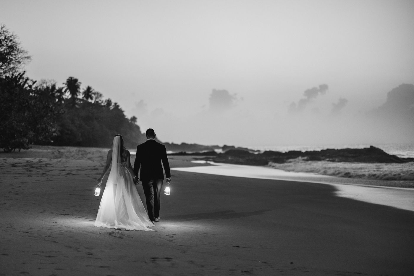 Black and white photo of a bride and groom walking on a beach at dusk, each holding a lantern.