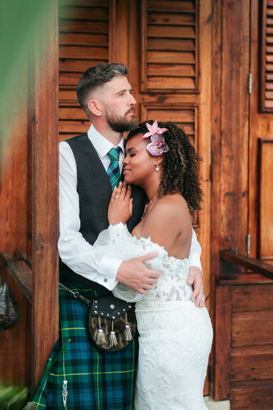 Interracial couple in wedding attire, groom in Scottish tartan kilt embracing bride in white lace gown by wooden doors.