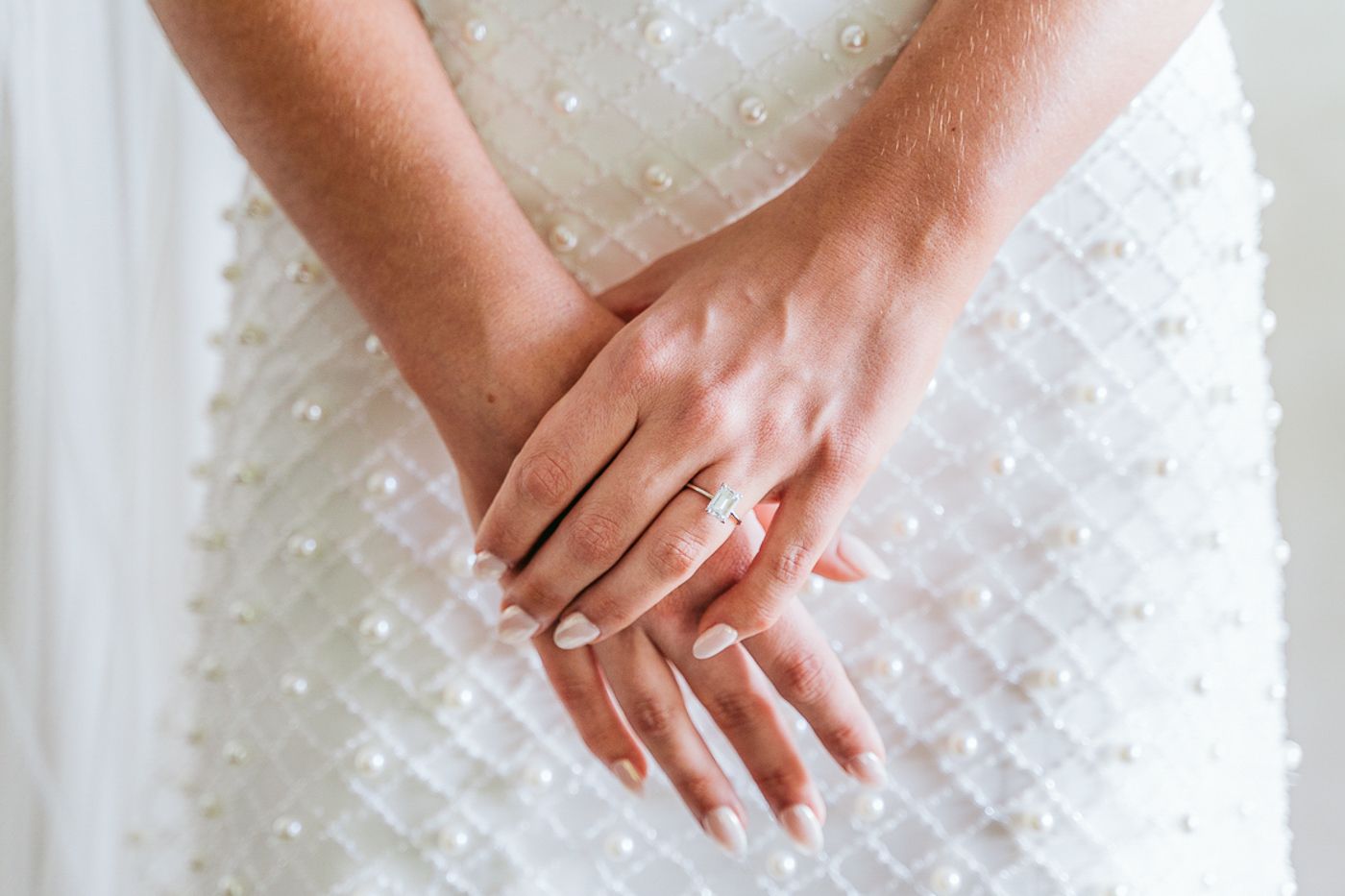 Close-up of bride's hands showing diamond engagement ring with French manicure against pearl-embellished white wedding dress.