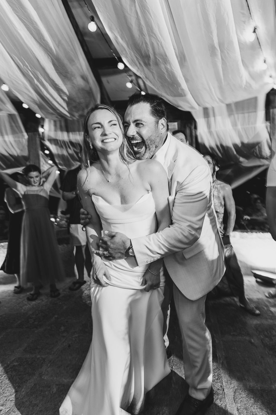 Black and white photo of joyful bride and groom embracing and laughing on wedding dance floor with draped fabric backdrop.