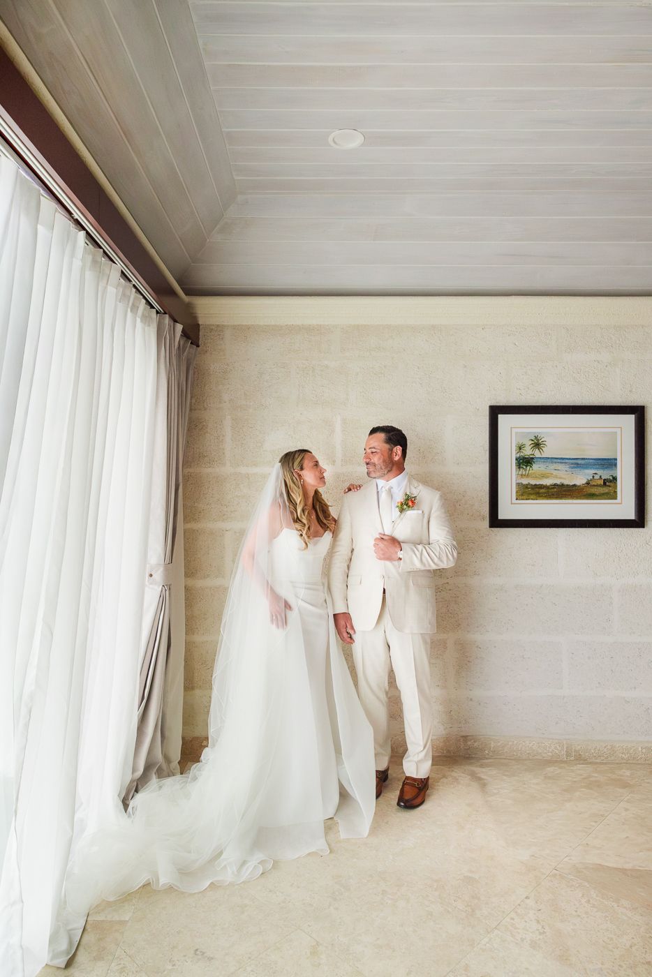 Bride in white gown and veil stands with groom in cream suit, sharing a joyful moment in an elegant stone-walled room.