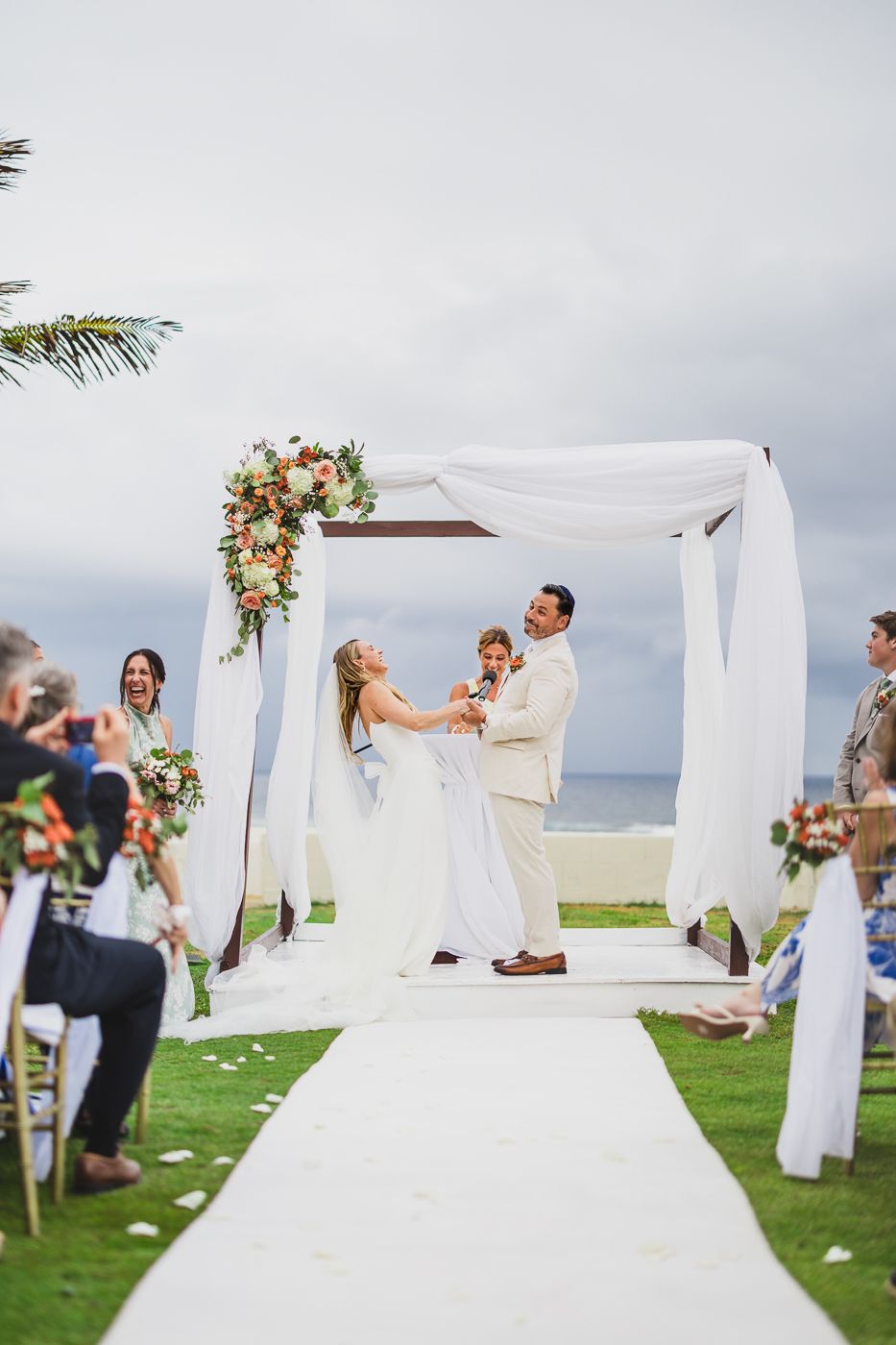 Bride and groom celebrate at outdoor beach wedding ceremony under white draped arch with colorful floral arrangements.