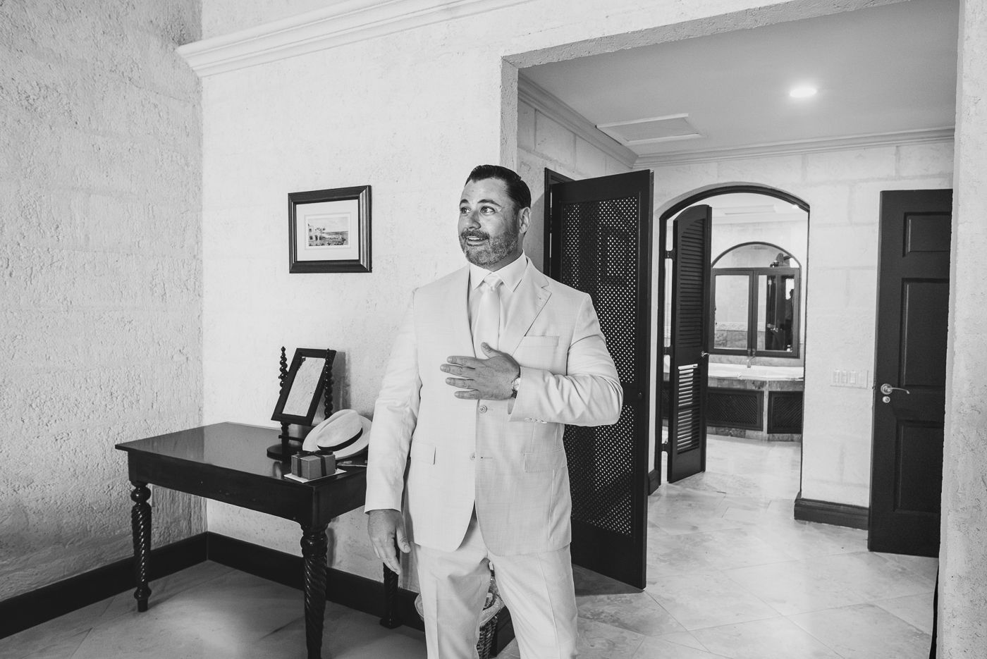 Groom in a light suit adjusting his tie in an elegant room before the wedding ceremony.