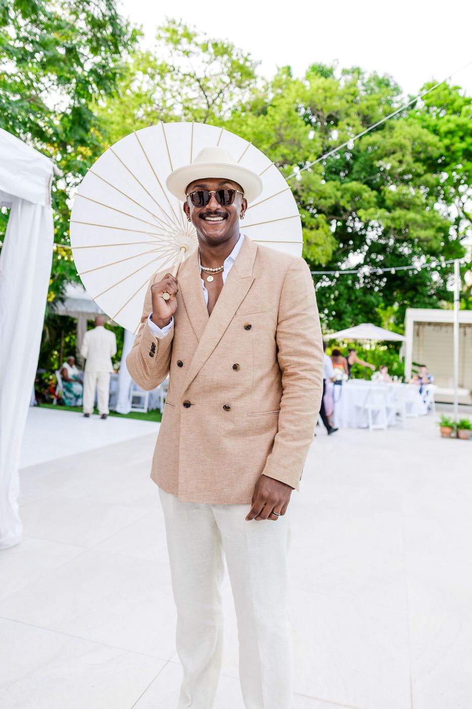 Stylish guest in a beige blazer holds a decorative parasol at an outdoor wedding reception.