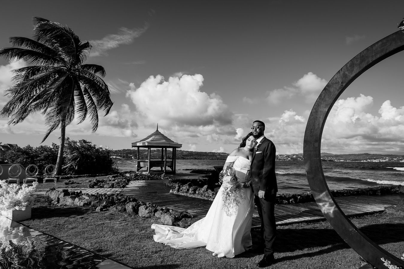 Black and white panoramic view of a beach wedding ceremony with palm trees and gazebo.