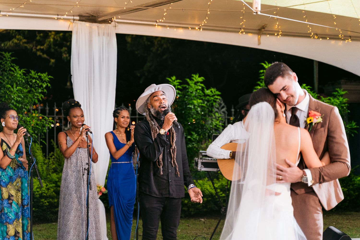 Wedding party celebrates and dances during outdoor reception under string lights and white fabric draping.