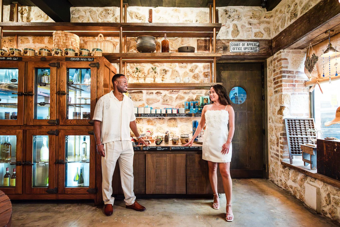 A couple poses in all-white outfits inside a rustic restaurant with vintage decor.