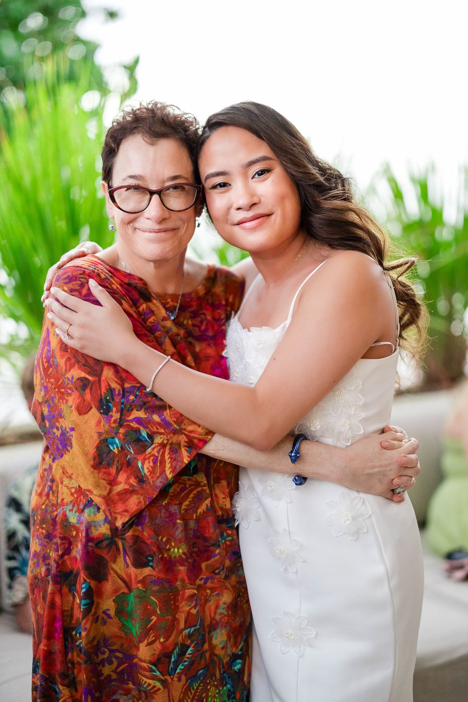 Two people share a warm embrace at a tropical venue, one wearing a colorful floral dress.