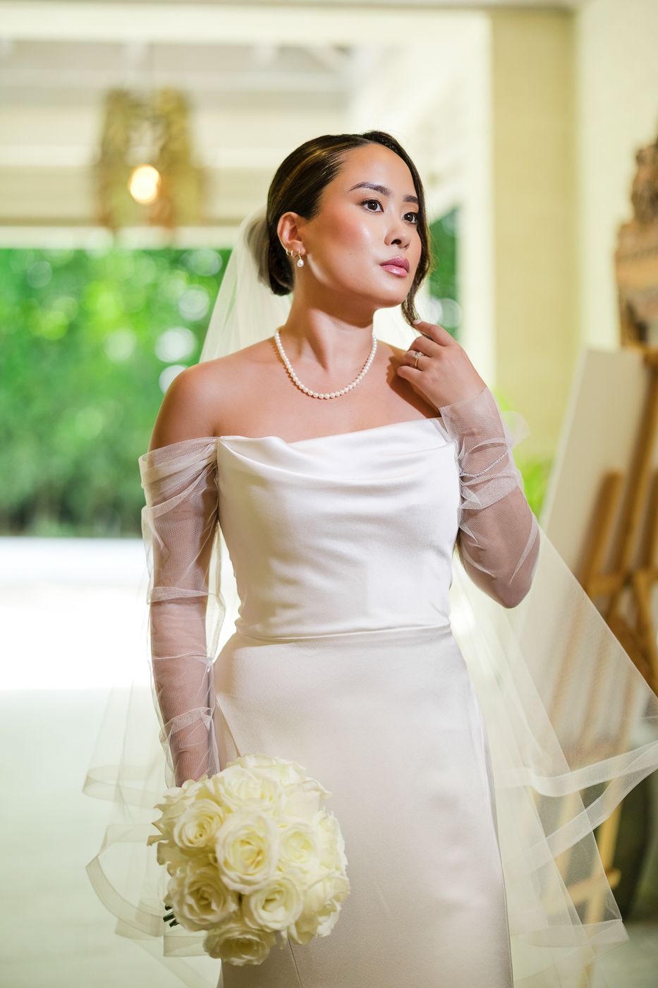 Bride in elegant off-shoulder gown holds white rose bouquet while posing in natural light.