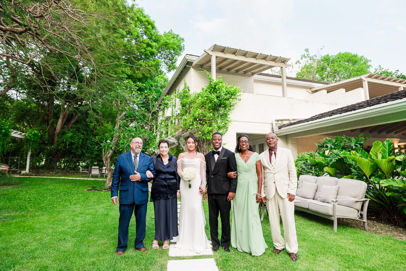 Wedding party gathered on landscaped lawn with elegant white architectural backdrop.