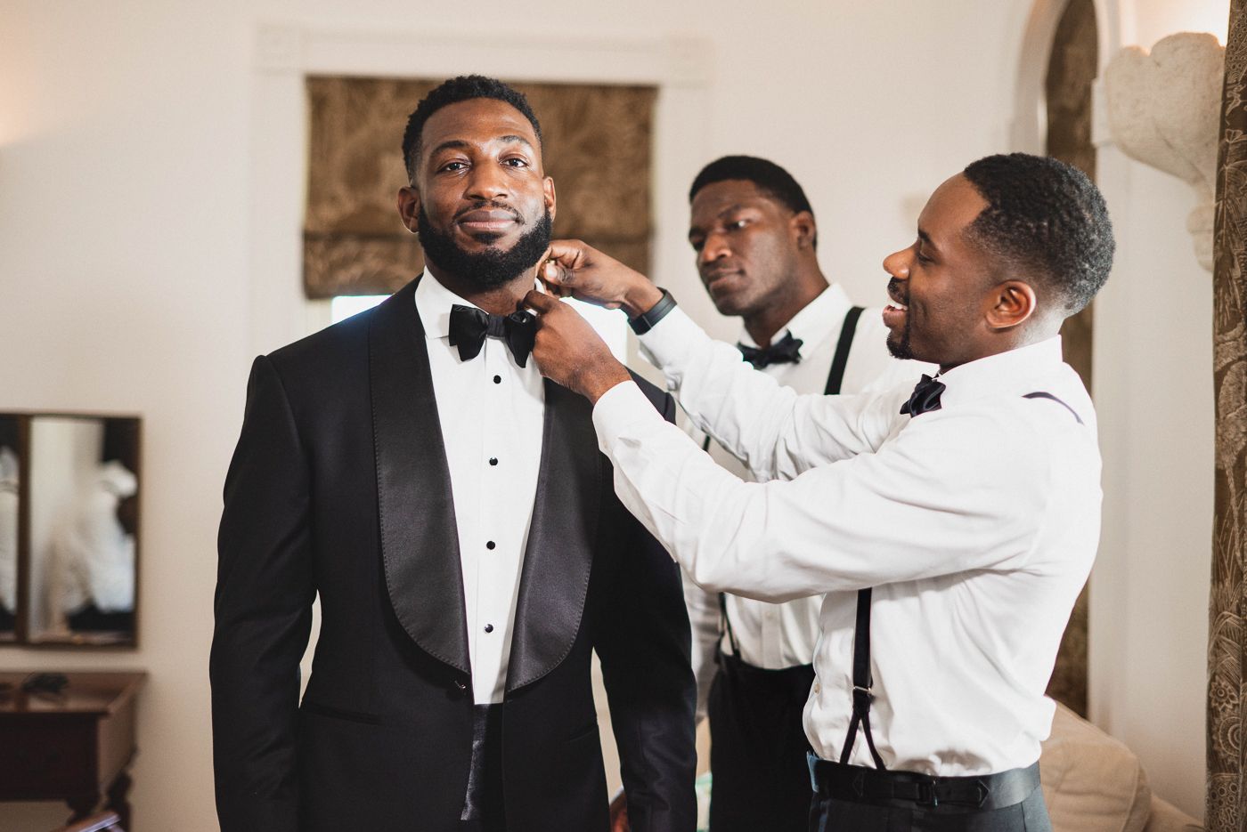 Groomsmen help adjust bowtie on groom's tuxedo during wedding preparations.
