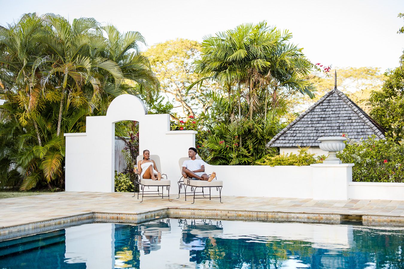 Two people relaxing on lounge chairs beside a tropical pool with lush palm trees and white villa walls.