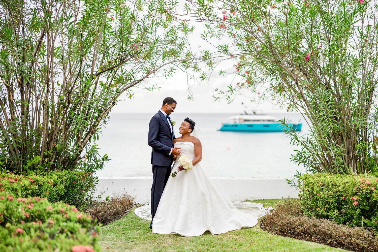 Bride and groom share a romantic moment under a lush green arch with a waterfront view on their wedding day.
