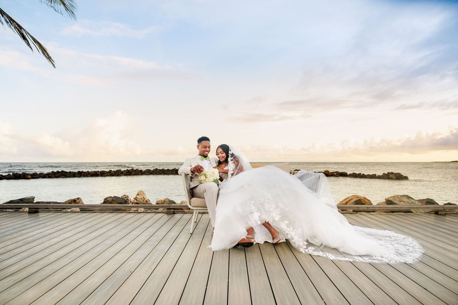 Bride and groom pose on a wooden deck by the ocean at sunset, her white gown trailing beautifully.