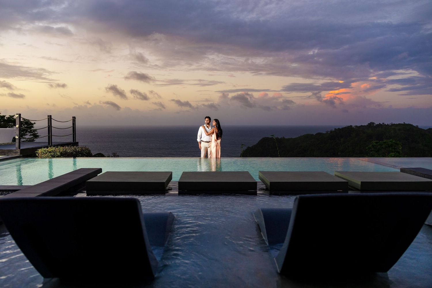 Couple embracing at infinity pool edge during dramatic sunset with ocean views and lounge chairs in foreground.