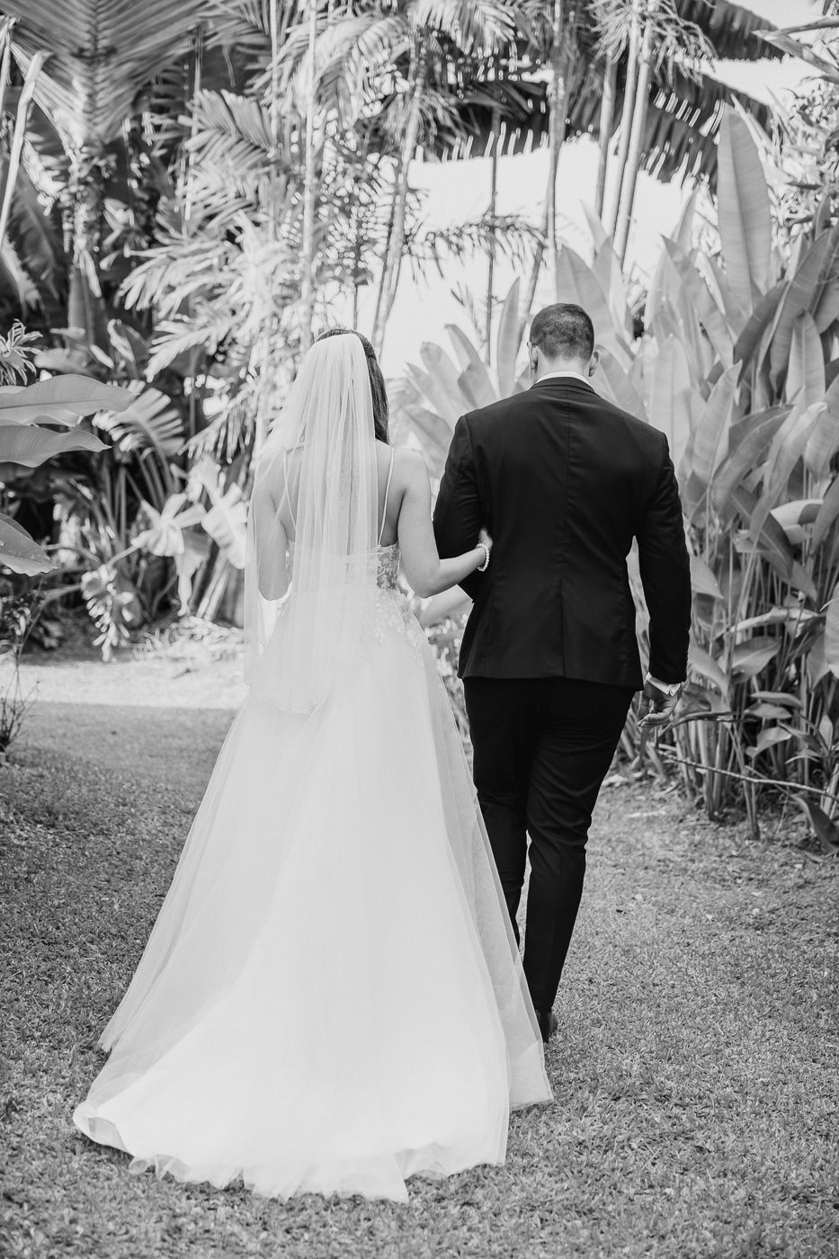 Black and white photo of a bride and groom walking hand in hand through a tropical garden on their wedding day.